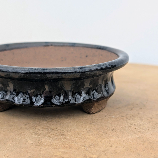 An artisan handmade ceramic bonsai pot by Brandon Besserer, featuring a low, round profile with a deep, glossy charcoal-black glaze. The exterior showcases a decorative scalloped texture around the base where the glaze thins to reveal silvery-blue iridescent highlights. The wide, rounded rim opens to a contrasting unglazed, reddish-brown clay interior. This small pot sits on textured unglazed cloud feet atop a light wood surface against a plain white background.