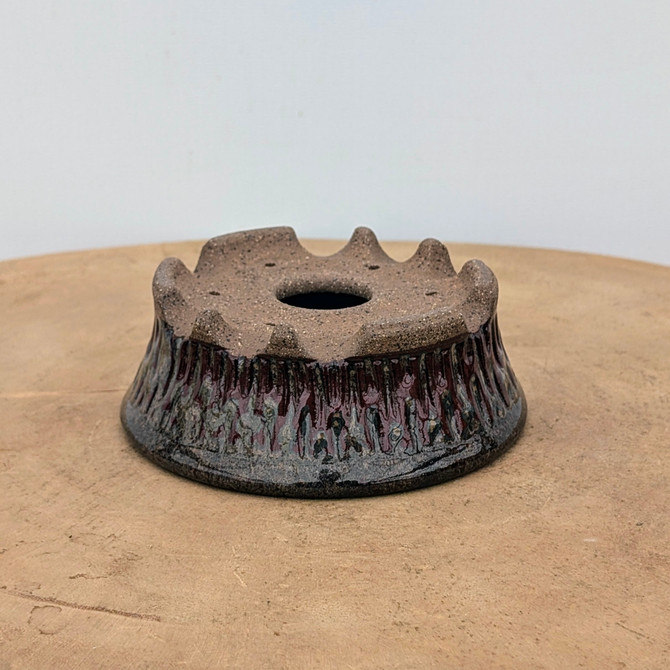 A detailed view of a handmade ceramic bonsai accent pot, identified as "No. 7" by artist Brandon Besserer. The pot features a unique, crown-like architectural shape with a wide, stable base that flares slightly inward before blooming out into a scalloped, notched rim. 

The flat top surface of the pot is unglazed, revealing a rustic, speckled sandy-brown clay texture. This top surface includes a large central drainage hole and several smaller holes for securing plants with wire. 

The exterior sides are textured with vertical ridges that create a fluted appearance. A dramatic, layered glaze covers these ridges, starting with a deep oxblood red or burgundy at the top that drips down into a mottled dark charcoal grey and metallic silver finish near the base. The pot sits on a light-colored, natural wood turntable against a clean, minimalist white background, emphasizing its artisanal craftsmanship and earthy aesthetic.