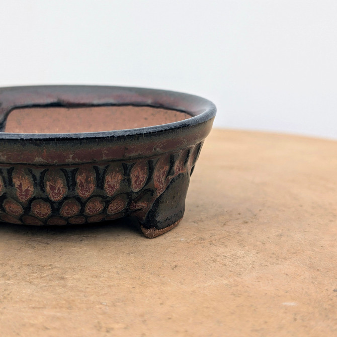 A detailed, side-profile close-up of a handmade ceramic bonsai pot by artist Brandon Besserer, specifically piece No. 11. The shallow pot is crafted with a textured, earthy finish, featuring a blend of dark charcoal and deep rust-red glazes. Its most striking feature is an intricate, repeating pattern of oval or scale-like shapes circling the base, each filled with a delicate, marbled pinkish-red design. The pot has a slightly flared rim and sits on small, sturdy feet. The photograph captures the pot resting on a light brown, textured surface against a stark white background, emphasizing the fine details and artisanal quality of the bonsai container.