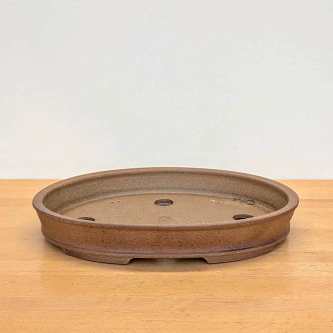 A detailed view of a shallow, handmade oval bonsai pot by artist Jack Hoover. The pot is crafted from unglazed brown clay with a grainy, textured finish, giving it a natural and rustic appearance. It features an elegant oval shape with a decorative braided-pattern band encircling the exterior just below the upper rim. The base is slightly raised with subtle, integrated feet. Inside, the pot has a flat bottom with three circular drainage holes for optimal water management and a faint artist's seal visible in the center. The pot is centered on a light-toned wooden tabletop against a plain, off-white background, highlighting its artisanal craftsmanship.