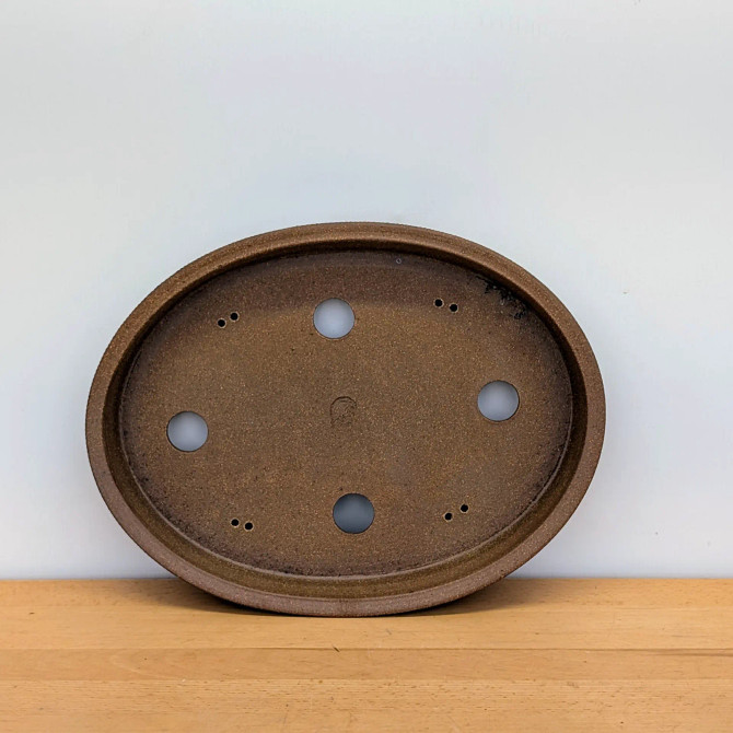 An overhead view shows a handcrafted, oval bonsai pot by artist Jack Hoover, showcasing its functional design and rustic aesthetic. The ceramic pot is finished in a warm, earthy brown with a finely textured, matte surface that gives it a natural look. 

Inside the pot's shallow basin, four large circular drainage holes are arranged in a diamond pattern to ensure proper water flow. Between these larger openings are four pairs of smaller, pin-sized holes designed for securing bonsai wire to stabilize a tree. In the very center of the textured base, a small, circular potter's mark is subtly stamped. The pot is shown leaning slightly forward on a light-toned wooden shelf against a plain white background, highlighting its clean lines and handcrafted quality.