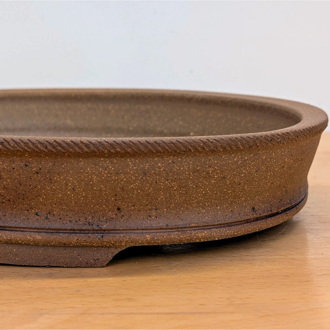 A detailed close-up side profile of a handmade ceramic bonsai pot by artist Jack Hoover. The unglazed pot is crafted from a rich, reddish-brown clay featuring a speckled texture with small dark brown and black mineral deposits throughout. A decorative, rope-like braided pattern is meticulously carved into the top outer rim, adding a tactile, artisanal detail. Near the base, two fine horizontal grooves encircle the pot, leading down to a recessed "cloud" foot design that elevates the container slightly off the light-colored wooden surface it sits on. The background is a clean, neutral off-white, which emphasizes the earthy tones and fine craftsmanship of the pottery.