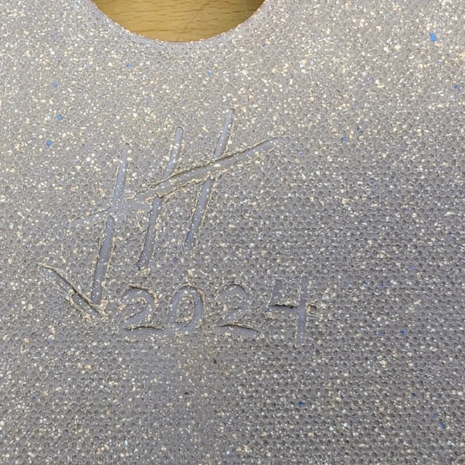 A close-up photograph of the underside of a handmade ceramic bonsai pot by artist Jack Hoover. The unglazed clay has a gritty, granular texture with a base color of tan and light gray, peppered with fine dark spots and occasional small blue mineral inclusions. 

Centrally located is a hand-incised artist's mark or monogram, consisting of three vertical carved lines with a single diagonal stroke slashing across them from the bottom left toward the top right. Below this stylized signature, the year "2024" is clearly inscribed into the clay. At the top edge of the frame, the curve of a circular drainage hole is visible, revealing the light-colored wooden surface beneath the pot. This detail highlights the craftsmanship and individual character typical of custom bonsai pottery.