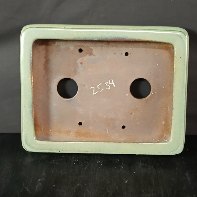 A top-down, overhead view of a 12-inch shallow rectangular Yixing ceramic bonsai pot, model number 2534. The pot features a thick, rounded rim finished in a smooth, light sage-green glaze. The interior is unglazed, showcasing the natural, earthy reddish-brown texture of the Yixing clay. At the center of the interior, the number '2534' is handwritten in white. The pot is designed with functional features for bonsai cultivation, including two large circular drainage holes on the left and right, and four smaller wiring holes positioned near each of the four rounded corners. The pot is presented on a dark surface against a neutral black backdrop, emphasizing its clean lines and subtle green glaze.