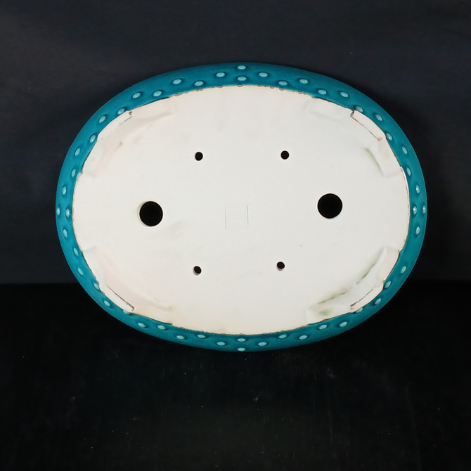 A detailed bottom-up view of a 12-inch oval Yixing ceramic bonsai pot (model No. 2512D), highlighting its functional and decorative features. The pot's exterior rim is finished in a vibrant, glossy turquoise glaze and is adorned with a row of raised, circular rivets. The unglazed base reveals a smooth, off-white ceramic clay body. For proper plant care, the bottom includes two large circular drainage holes and four smaller holes designed for bonsai wire to secure the tree. Four low, integrated feet provide elevation for air circulation. A square maker's mark or seal is embossed in the center of the base. The entire object is set against a solid black background, emphasizing its color, shape, and craftsmanship.