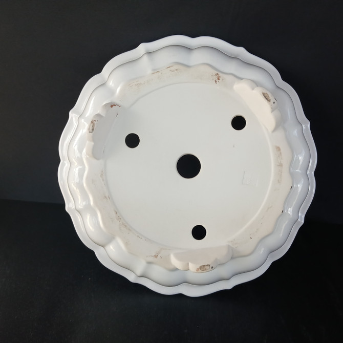 A high-angle view showcasing the underside of a 17-inch white round glazed Yixing bonsai pot. The pot features a beautiful scalloped or fluted rim, giving it a decorative, petal-like appearance. The glossy white ceramic base has four drainage holes: a large central one surrounded by three smaller ones, ensuring excellent water flow for a bonsai tree. Four small, rounded feet are built into the design to lift the pot slightly off the surface. These feet and the contact ring are unglazed, showing some natural clay color and minor wear. The clean, minimalist aesthetic of the white glaze stands out sharply against the plain black background, highlighting the pot's structure and functional details.