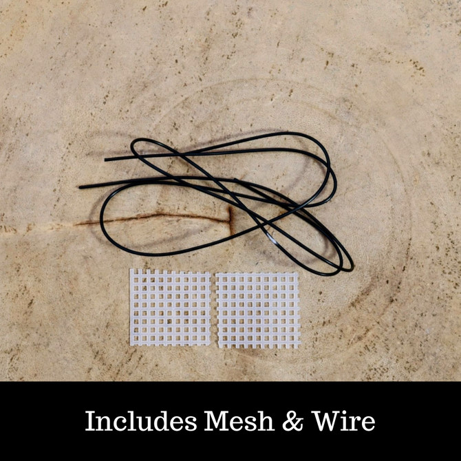 A top-down photograph shows a bundle of thin black bonsai wire and two small squares of white plastic mesh drainage screens. The wire is loosely coiled in an irregular loop, resting above the two identical mesh squares which are placed side-by-side. These items are laid out on a light-tan, textured surface that resembles the cross-section of a piece of wood or bark, featuring faint concentric rings and a small horizontal crack. 

At the bottom of the image, a solid black banner contains the white serif text, "Includes Mesh & Wire." These components are essential bonsai pot accessories; the mesh squares are used to cover drainage holes to prevent soil from washing out, while the wire is used to secure the bonsai tree within its pot. This set is specifically included with the 12-inch glazed oval Yixing bonsai pot.