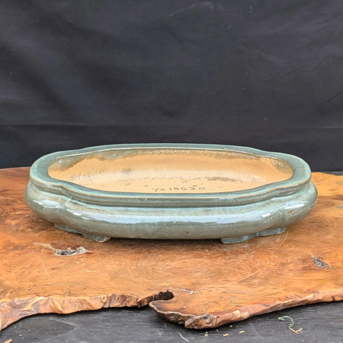 An elegant, shallow Yixing bonsai planter sits atop a polished, irregular wood slab. This oval-shaped pot, identified as SKU 1862G, features a classic lobed or scalloped rim and is finished in a glossy, muted celadon glaze with subtle variations in its greenish-grey tone. The planter rests on small, integrated cloud feet. Inside, the pot remains unglazed, revealing the natural buff-colored clay body where the code 'yx 1862 G' is handwritten in black ink. The piece is displayed against a dark, solid backdrop, highlighting the smooth glaze and the rich grain of the wooden base.