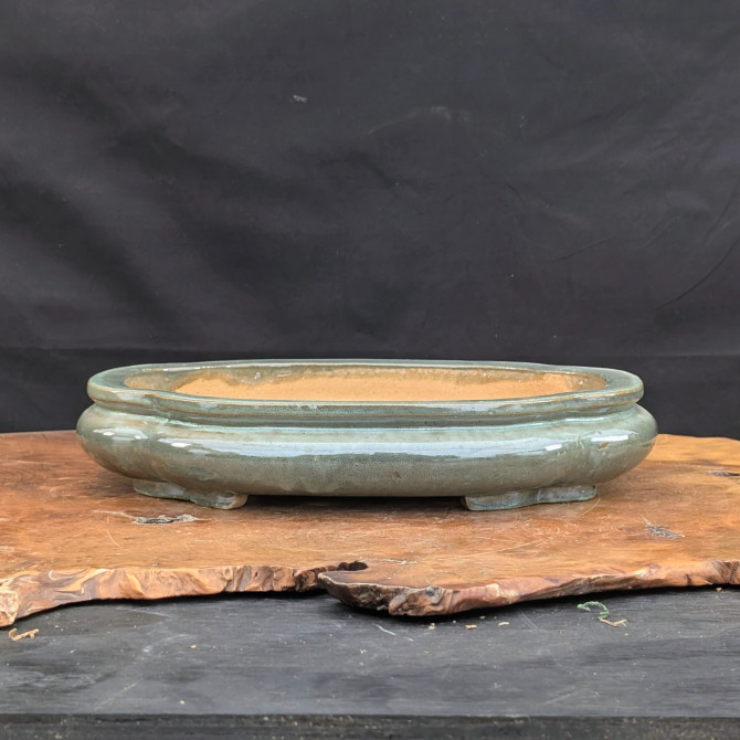 A shallow, oval-shaped Yixing ceramic bonsai pot sits centered on a rustic wood slab. The pot features a smooth, glossy glaze in a muted grayish-green or celadon color, with subtle variations and highlights reflecting from its curved surface. Its design includes a slightly rounded body, a thin defined rim, and four small, integrated feet that lift it slightly off the surface. The interior of the pot is unglazed, revealing the natural, warm tan-orange color of the clay. The planter is displayed on a thick, dark-grained wooden base with a natural live edge, which rests on a dark foreground against a solid black fabric backdrop, emphasizing the bonsai pot's elegant form and earthy color palette.