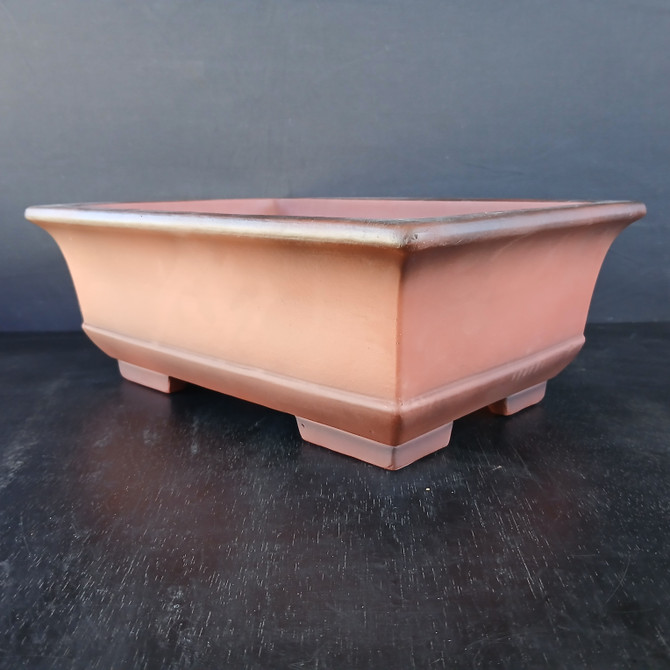 An image featuring a large, 16-inch unglazed rectangular Yixing bonsai pot (model YX2283-1). The pot is crafted from traditional reddish-brown clay with a smooth, matte finish and subtle, darker smoky-toned accents highlighting the flared upper rim and the base. Its sturdy design features slightly tapered sides and is elevated by four block-shaped corner feet that ensure proper drainage and airflow for a bonsai tree. The container is photographed against a dark, neutral background on a black surface with a subtle wood grain, making the warm, earthy tones and professional structure of the Yixing clay stand out.