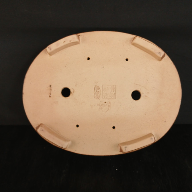A detailed top-down view of the underside of a 12-inch oval Yixing bonsai pot, showcasing its light beige, unglazed ceramic surface. The pot features four sturdy, rectangular feet at the quadrants for elevation and stability. 

Two prominent circular drainage holes are located along the central horizontal axis, accompanied by four smaller wire holes arranged symmetrically in a rectangular pattern for securing a bonsai tree to the container. At the very center, an authentic manufacturer's chop mark or seal with Chinese characters is embossed into the clay, indicating its craftsmanship. The entire bottom of the pot is set against a dark, non-reflective background, highlighting its clean lines and the matte texture of the Yixing clay.