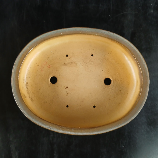 A top-down view of a 12-inch oval glazed Yixing bonsai pot, identified by the model number 2532H. The interior of the pot has a smooth, matte finish in a warm sand or light tan color, which contrasts with the dark grey glaze visible along the top rim. At the bottom of the pot, two large circular drainage holes are positioned on the left and right sides to ensure proper water runoff. Four smaller holes are arranged in a rectangular pattern around the center for threading bonsai wire to secure a tree in place. On the left side of the interior floor, the model number '2532 H' is faintly stamped in red ink. The pot is placed against a solid black, textured background that emphasizes its oval shape and functional design.