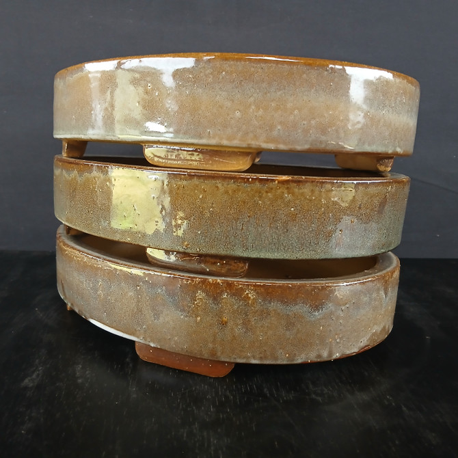 A stack of three 12-inch glazed oval Yixing bonsai pots is presented against a dark, minimalist background. The pots are stacked vertically, showcasing their uniform, shallow elliptical shape and low profile. Each pot features a rich, high-gloss glaze with a mottled, earthy color palette of amber, tan, and dark brown, with subtle hints of olive green and a speckled "oil spot" texture.

The glaze is reflective, catching the light and highlighting the smooth, curved edges of the ceramic. Small, integrated rectangular feet are visible at the base of each pot, designed to provide elevation for proper drainage and airflow. The stack sits on a dark, slightly reflective surface, which emphasizes the warm tones and artisanal quality of the Yixing clay. The overall composition highlights the natural variation in the glazing process, making each pot appear unique while maintaining a cohesive aesthetic.