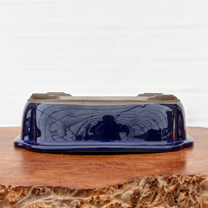 An upside-down view of a rectangular Japanese Tokoname bonsai pot, showcasing its signature deep blue Namako glaze and unglazed bottom. The glossy, cobalt blue finish is highly reflective, capturing distorted light and shadows, while the top-facing base reveals the natural, matte brown clay and four distinct integral feet. The pot rests on a textured, live-edge wooden display stand against a stark white background, emphasizing the elegant contrast between the polished ceramic finish and the rustic, organic wood surface.