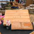 A high-angle, professional product shot displays the Tinyroots Ultimate Stainless Steel Bonsai Tool Kit components, highlighting a blend of craftsmanship and utility. The centerpiece is a large, light-colored bamboo tool chest with the "Tinyroots" brand name and Japanese calligraphy for "Bonsai Tools" elegantly engraved on the lid. 

To the left of the chest stands a small, upright box of Tinyroots Choji Oil, marked as a rust prevention solution for keeping stainless steel tools in peak condition. In the foreground, a folded, light brown suede tool roll is laid out, featuring a stitched leather patch with the Tinyroots logo. 

The entire set is positioned on a black, textured workbench. The background, softly blurred to keep focus on the product, reveals the setting of a bonsai nursery or workshop, with stacks of colorful glazed ceramic bonsai pots in various sizes and earth-toned bags of specialized bonsai soil and substrate piled nearby. This composition emphasizes the high-quality, professional nature of the bonsai maintenance equipment.