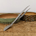 A professional carbon steel bonsai combination tool, featuring a pair of long-reach tweezers and a curved spatula, is displayed on a weathered wooden tabletop. The tool is positioned diagonally across the frame; its broad, flat spatula end rests on the light brown, scratched wood in the lower-left corner, while the long, slender tweezer arms extend upwards toward the top-right. The tweezers feature fine, serrated tips for precision work.

The tool is propped against two shallow, handmade ceramic bonsai pots. The pot on the left has a simple, speckled sage-green and earthy-brown glaze. The pot on the right is more ornate, featuring a deeply textured, carved exterior with a complex glaze of teal, blue, and dark brown. The background is a clean, neutral off-white wall, which makes the metallic grey of the carbon steel tool and the rich textures of the ceramic pots stand out. This image highlights a versatile piece of equipment essential for delicate bonsai maintenance tasks like repotting, weeding, and soil grooming.