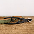 An high-angle, close-up photograph features a pair of professional-grade Tinyroots carbon steel bonsai concave cutters lying horizontally on a weathered wooden workbench. The long, elegant black handles extend to the left, with the brand name "Tinyroots" clearly embossed into the metal near the central pivot rivet. To the right, the sharp, heavy-duty concave blades are slightly open, revealing their bright, polished steel cutting edges. 

The tool is artfully positioned resting atop two small, artisan ceramic bonsai pots. One pot is a shallow, sage-green speckled dish, while the other features a deeply textured, earthy-toned surface with hints of blue and green glaze. The scene is set against a neutral, off-white background, emphasizing the sleek design and high-quality construction of this essential bonsai pruning tool, which is a key component of the Tinyroots Ultimate Carbon Steel Tool Kit.
