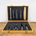 An open, light-colored bamboo wooden box displays a comprehensive 10-piece Tinyroots Ultimate Package carbon steel bonsai tool kit. The set features an array of professional-grade tools, including various cutters, pruning shears, bud shears, a root rake with a spatula end, tweezers, and a folding saw with a light wood handle. 

Each tool is neatly organized and securely held in place within custom-cut black foam inserts in both the lid and the base of the case. The box itself has elegant brass-colored metal hinges and latches and is photographed on a rustic, weathered wooden tabletop against a clean white background. This presentation highlights the high-quality craftsmanship of the carbon steel tools designed for precise bonsai tree care and maintenance.