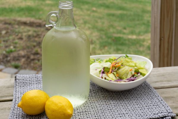 Glass jug bottle filled with fresh lemonade next to two lemons and a salad.