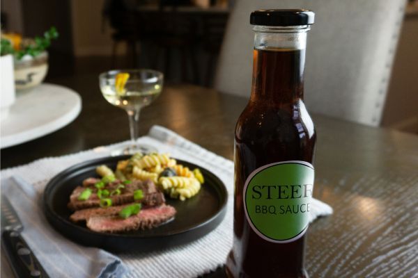 Glass bottle of Steer BBQ sauce on a table next to a plate of sliced steak and pasta.
