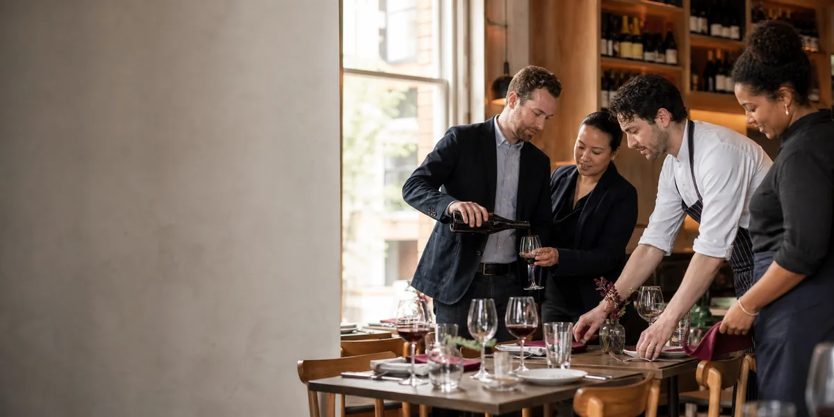 A wine professional pouring wine as a restaurant team prepares a dining table