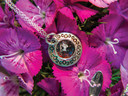 A close-up shot of a sterling silver tea cup pendant nestled among bright magenta dianthus flowers. The miniature cup is filled with real black and rose tea leaves encased in resin. The saucer beneath it features a swirling engraved pattern, and a delicate silver chain loops gracefully from the top.