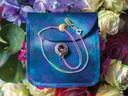 A top-down styled display featuring the tea cup pendant and chain neatly arranged on a blue velvet snap pouch, surrounded by a colorful ring of flowers. The scene feels magical and gift-ready, highlighting the craftsmanship and presentation.