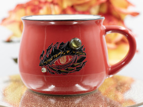 Red ceramic cauldron mug with matte gold "Dragon eye" design, black rim, white interior, and two Labradorite gemstones, front view on white background. Red ceramic cauldron mug with matte gold "Dragon eye" design, black rim, white interior, and two Labradorite gemstones, front view on white background.