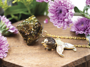 Flat lay of Water Nymph pendulum with mermaid tail charm, smoky quartz, and gold-plated chain, styled with pink blossoms.