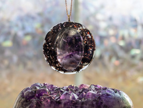 A close-up shot of a round amethyst pendant suspended on a gold chain. The centerpiece is a polished, domed amethyst cabochon surrounded by a ring of shimmering black tea leaves and copper flecks encased in resin. The pendant is photographed above an amethyst crystal cluster, with soft iridescent bokeh in the background.