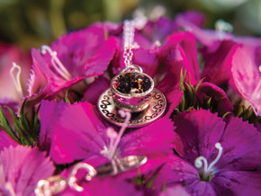 A side angle of the sterling silver tea cup pendant resting on vibrant pink flowers. The resin-filled cup glows softly in the light, revealing textured tea leaves inside. The engraved saucer adds a whimsical, ornate touch to the tiny wearable tea cup.
