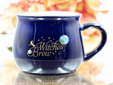 Royal blue ceramic cauldron mug with matte gold "Witches Brew" design, black rim, white interior, and two Labradorite gemstones, front view on white background.