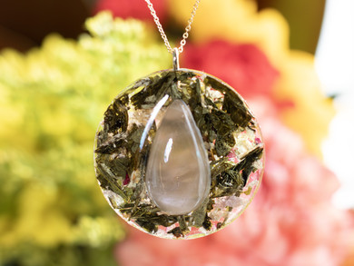 A close-up of a circular pendant featuring a teardrop-shaped rose quartz cabochon at the center. Surrounding the stone are suspended green tea leaves and tiny pale pink flower petals encased in clear resin. The background is filled with softly blurred yellow and pink flowers, creating a warm, dreamy glow.