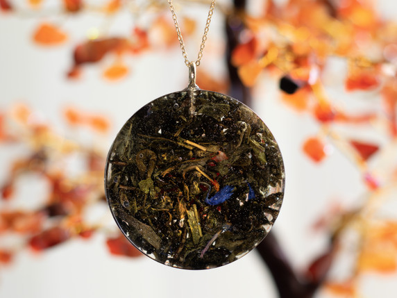 Back view of the pendant suspended against a softly blurred carnelian crystal tree. The rear of the pendant reveals embedded green tea leaves, flecks of gold, and a touch of blue flower petals sealed within the resin, giving it a natural, earthy texture.