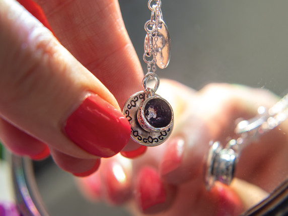 A hand with glossy red nails gently holds the tea cup pendant up to a mirror. The reflective surface captures a second view of the pendant, emphasizing the tiny sculpted details, the sterling silver shine, and the tea leaves suspended inside.
