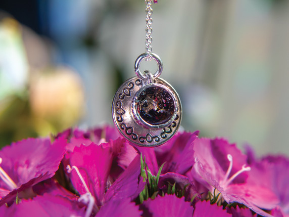 The tea cup pendant hangs suspended above a bed of rich fuchsia petals. The camera focuses on the front of the cup, allowing the shimmering tea-leaf resin and detailed silver etching on the saucer to stand out beautifully.