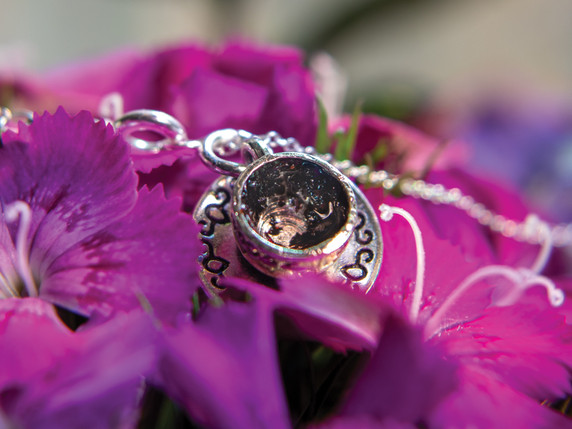 A close-up of the pendant resting among pink dianthus petals. The angle highlights the depth and glimmer inside the cup, along with the ornate etched pattern around the saucer. Soft natural light enhances the warm tones of the tea leaves.