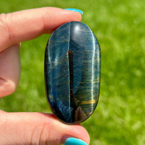 Blue Tigers Eye palm stone held between fingertips, a polished oval with deep blue chatoyant bands and a flash of gold