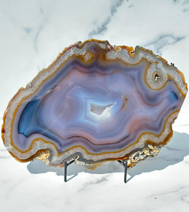 Large banded blue agate slab with quartz druzy center, front polished, displayed on a black metal stand against white marble