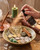 Hand misting Cinnamon Crystal Spray over a prosperity bowl with coins, folded bills, cinnamon sticks, and crystals beside a lit candle