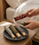 Lighting a rose dipped Palo Santo stick over a stone dish holding the blue sage, rosemary and lavender sticks