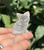 Clear Quartz sitting cat held on fingertips against green leaves, showing its milky translucent glow