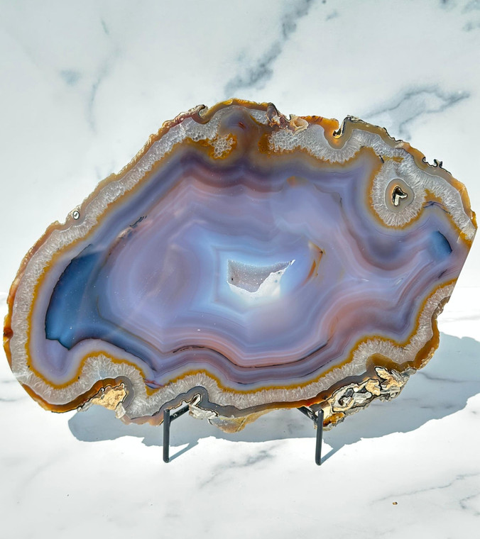 Large banded blue agate slab with quartz druzy center, front polished, displayed on a black metal stand against white marble