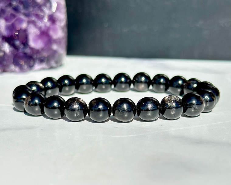 Hypersthene elastic bracelet with 8mm beads on white marble beside a purple amethyst crystal cluster