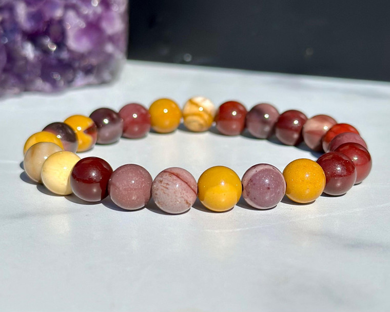 Mookaite elastic bracelet with 8mm mustard yellow, deep red, and cream beads resting beside a purple amethyst cluster
