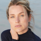 Woman in a navy turtle neck sweater, gazing at the camera with the ocean behind her.
