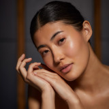 Woman with slicked-back hair resting her face against her hands in warm ambient lighting.