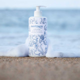 Phytomer Oleocreme  body milk bottle with blue floral pattern sitting on sand with ocean waves in the blurred background.