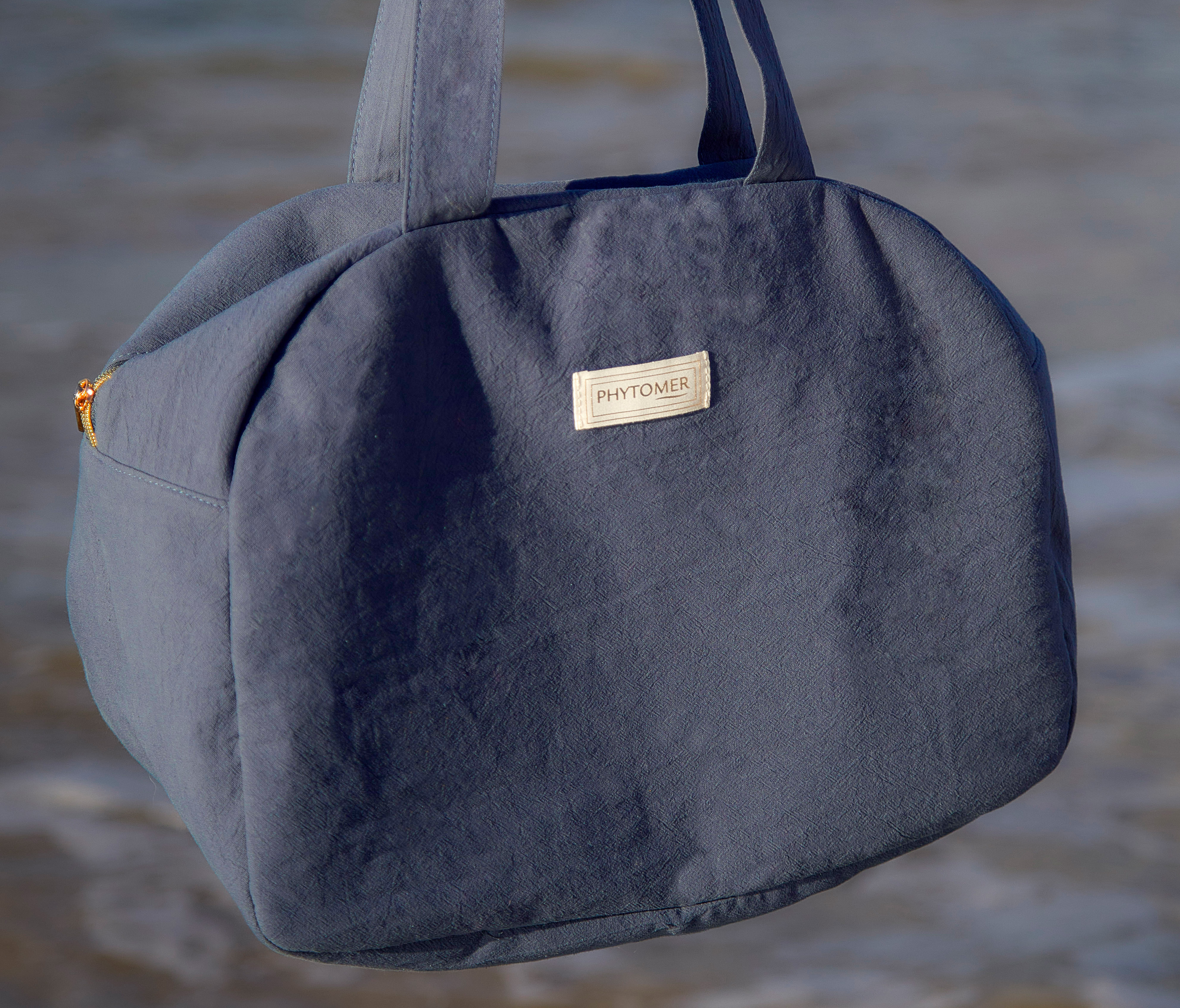 Phytomer branded blue tote bag held over the ocean.