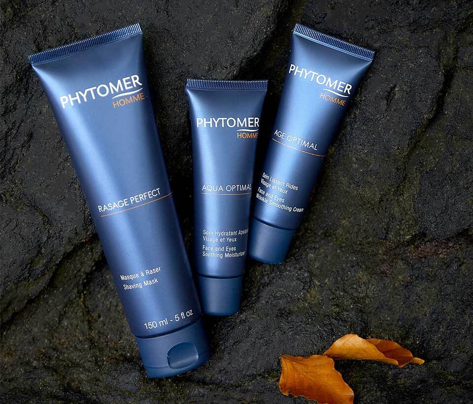 Rasage Perfect Shaving Mask 150 ml, Agua Optimal Face and Eyes Smoothing Moisturizer and the Age Optimal Face and Eyes Wrinkle Smoothing Cream lay side by side on a rock covered in leaves. 